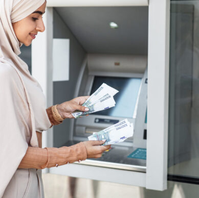 Debit and credit, money withdrawal, salary, money transfer, currency transfer outdoor. Business and work. Smiling young islamic lady in hijab counts dollars near atm machine in city, copy space
