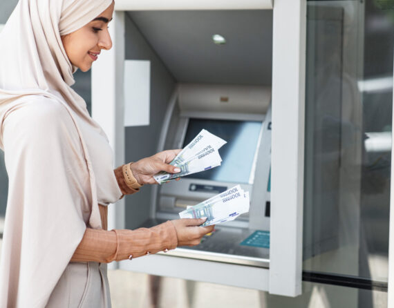 Debit and credit, money withdrawal, salary, money transfer, currency transfer outdoor. Business and work. Smiling young islamic lady in hijab counts dollars near atm machine in city, copy space
