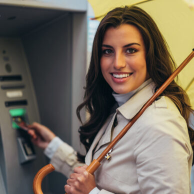 Beautiful young woman holding a yellow umbrella while using the atm machine