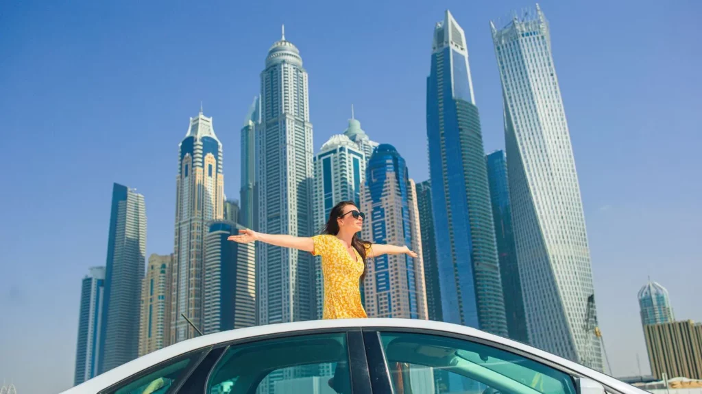 Louer une voiture à Dubaï : guide expatriés et touristes. 3 Touriste profitant de sa voiture de location à Dubaï devant les gratte ciel de la Marina